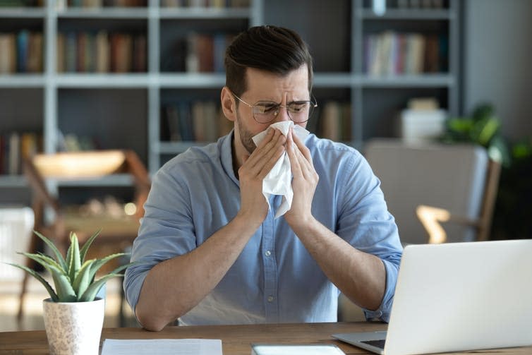 A man sitting at a desk blowing his nose with a tissue.