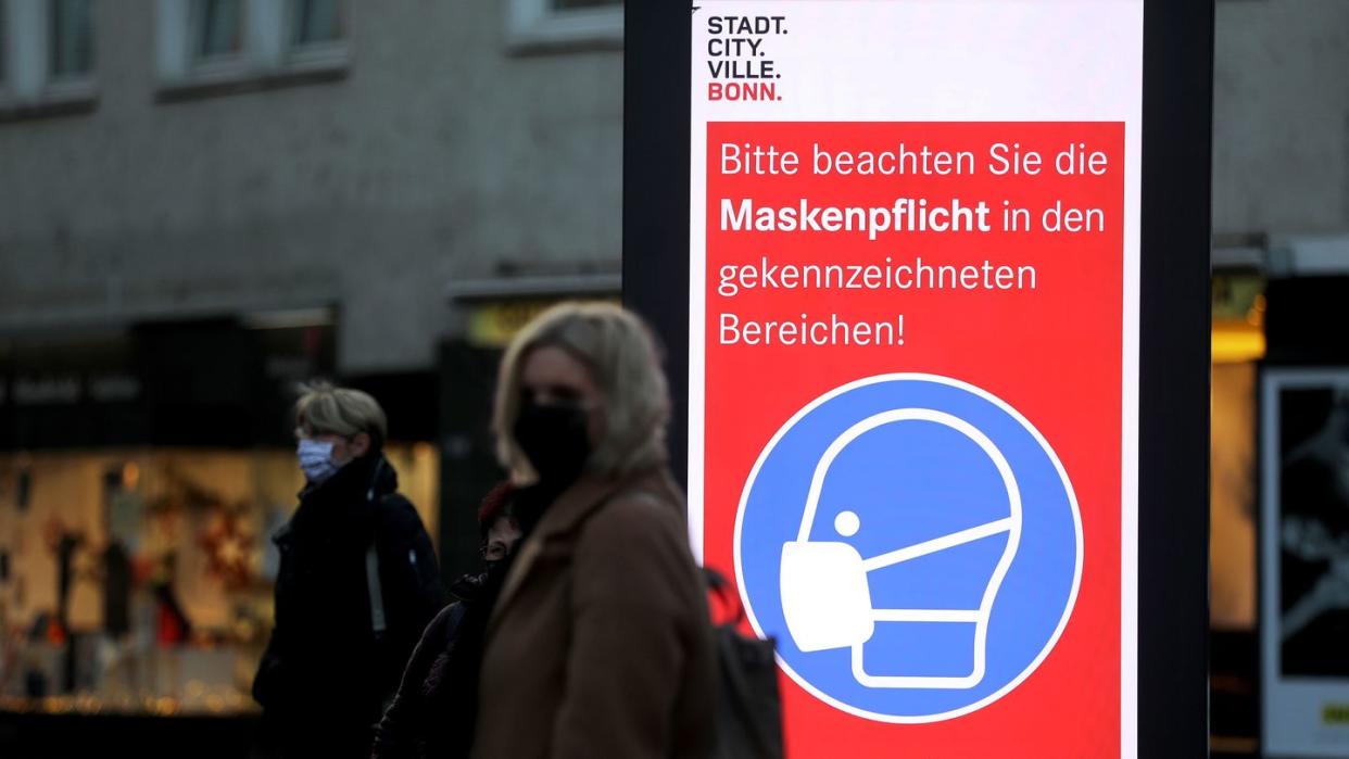 Frauen stehen neben einem Hinweisschild zur Maskenpflicht an einer Haltestelle in Bonn. Die Länder fordern vor den Beratungen mit Kanzlerin Merkel strengere Kontaktbeschränkungen, um die hohen Coronazahlen zu senken.