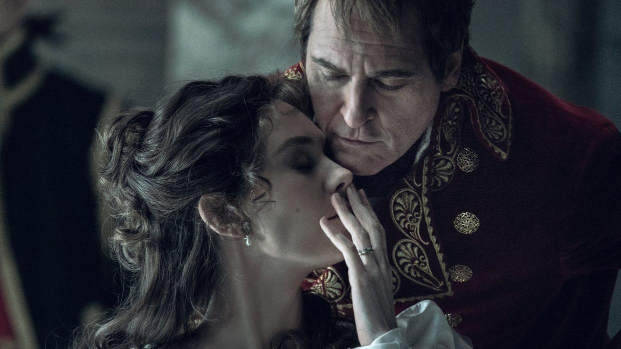 vanessa kirby and joaquin phoenix in character for napoleon, she has her head turned toward him and raises one hand over her mouth, he leans down to his head near hers, both have their eyes closed