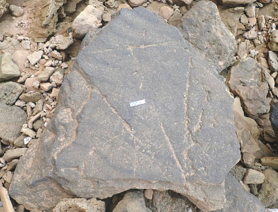 Una gran roca gris rodeada de piedras más pequeñas y granos de arena gruesos.  Hay tallas triangulares en la gran roca.