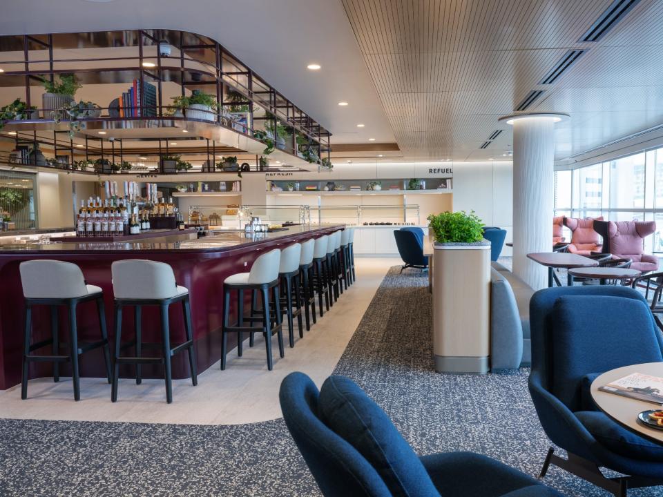A new airport lounge in Denver features a large open bar with red paneling on the left, and carpeted floors toward the right of the image with padded chairs and round tables for guests. A bank of windows on the right lets in natural light and gives guests a view of the tarmac.
