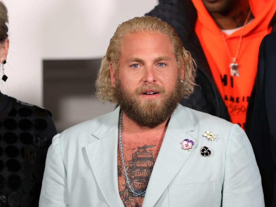 Jonah Hill in a light blue jacket showing off a chest tattoo