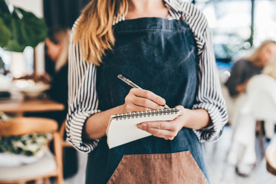 A woman wearing an apron over a striped shirt is writing in a notepad. The setting suggests she might be a waitress taking an order