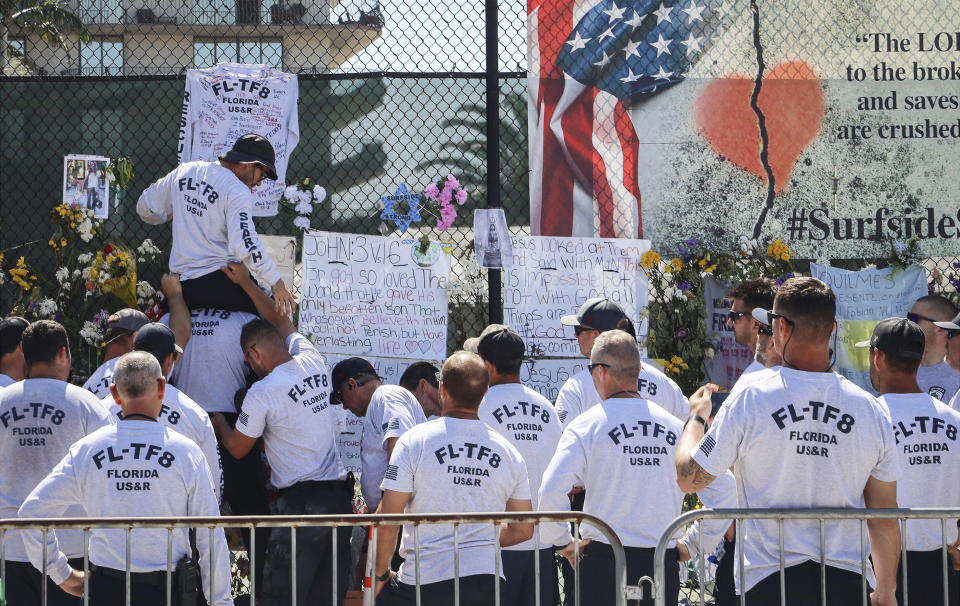 Members of the Florida Task Force 8 gather at the memorial site to pin an autograph shirt signed by team members on Sunday, July 4, 2021, in Surfside, Fla. Demolition specialists carefully bored holes to insert explosive charges into the precarious, still-standing portion of a collapsed South Florida condo building that will come down to open up new areas for rescue teams to search. A top Miami-Dade fire official said 80% of the drilling work was complete and the remaining structure could come down as soon as Sunday night. (Carl Juste/Miami Herald via AP)