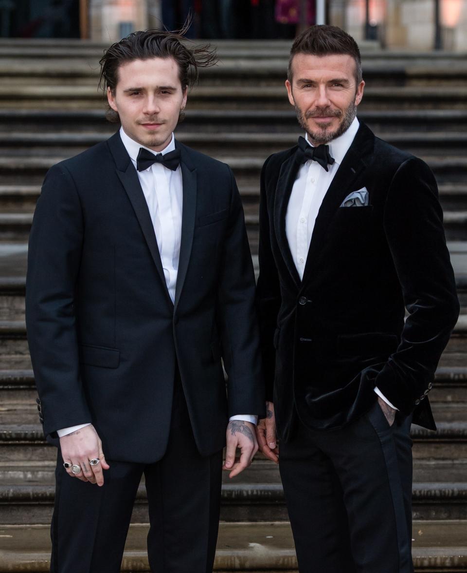 Brooklyn Beckham and David Beckham wear black suits as they're photographed on steps.