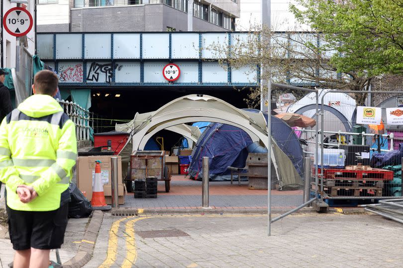 A security guard in a high-vis coat looks on at a cluster of tents in a fenced-off area of West Canal Wharf