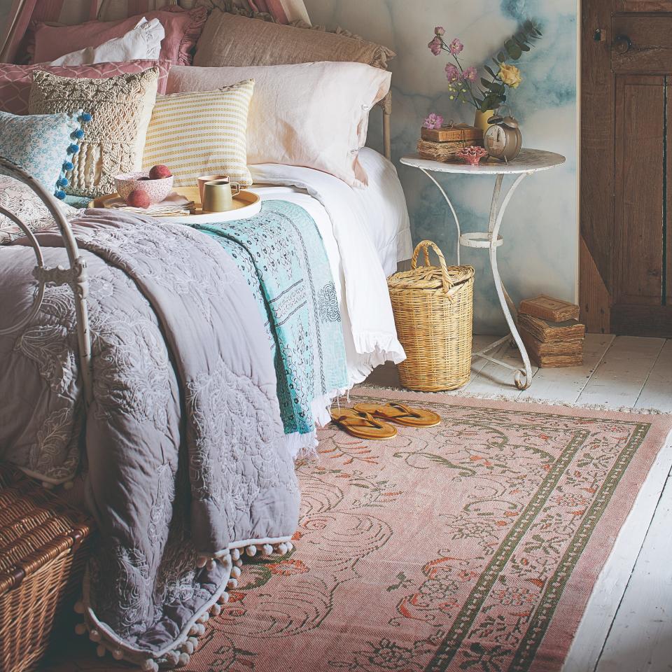 A bedroom with a pink pattertned rug and layered scatter cushions on the bed