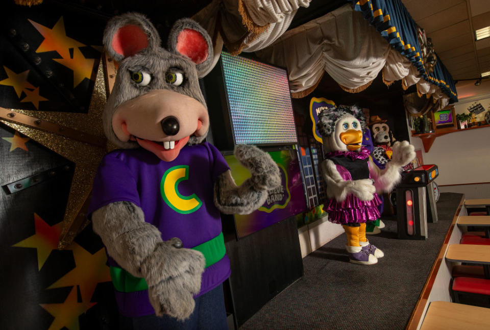 Chuck E. Cheese mascot in a purple shirt stands in foreground with animatronic band in background inside the venue