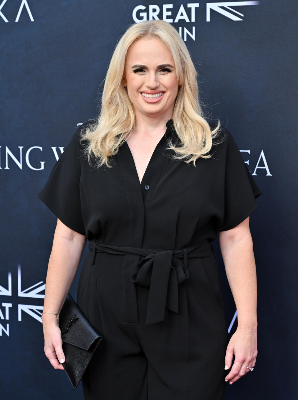 Rebel Wilson smiles while posing on the red carpet, wearing a black jumpsuit with a tie waist and holding a black clutch