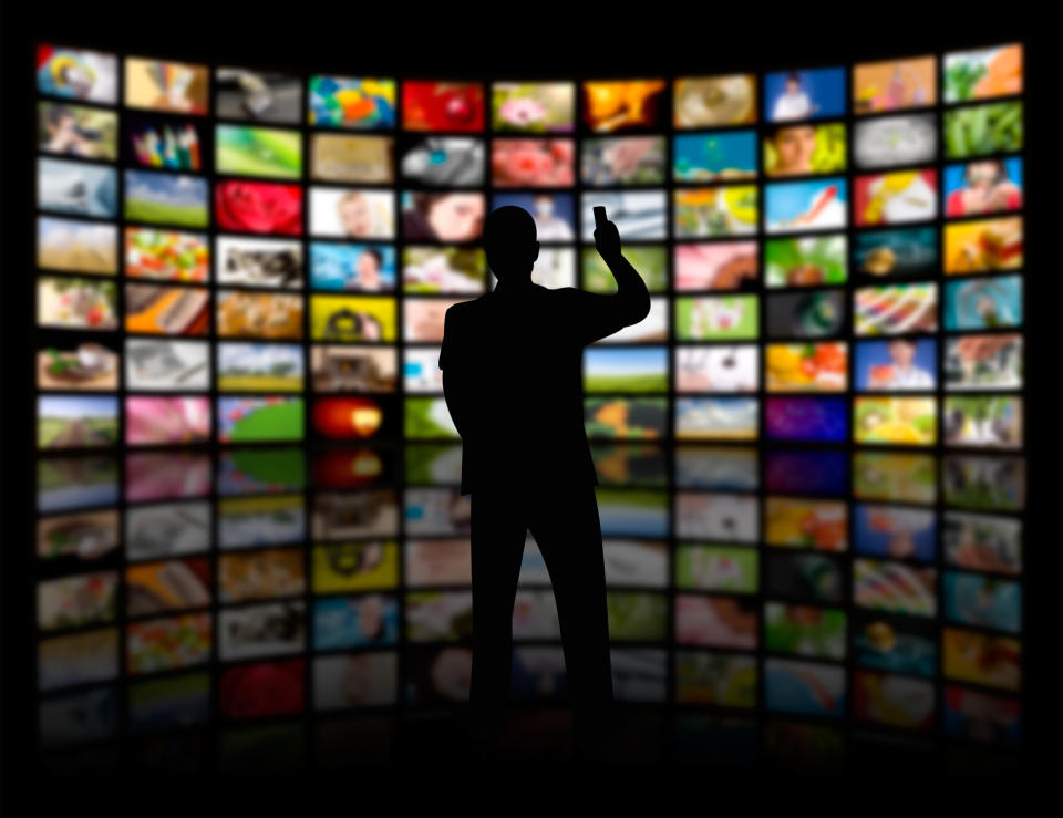 A man in silhouette with a remote facing a wall of dozens of TV screens.