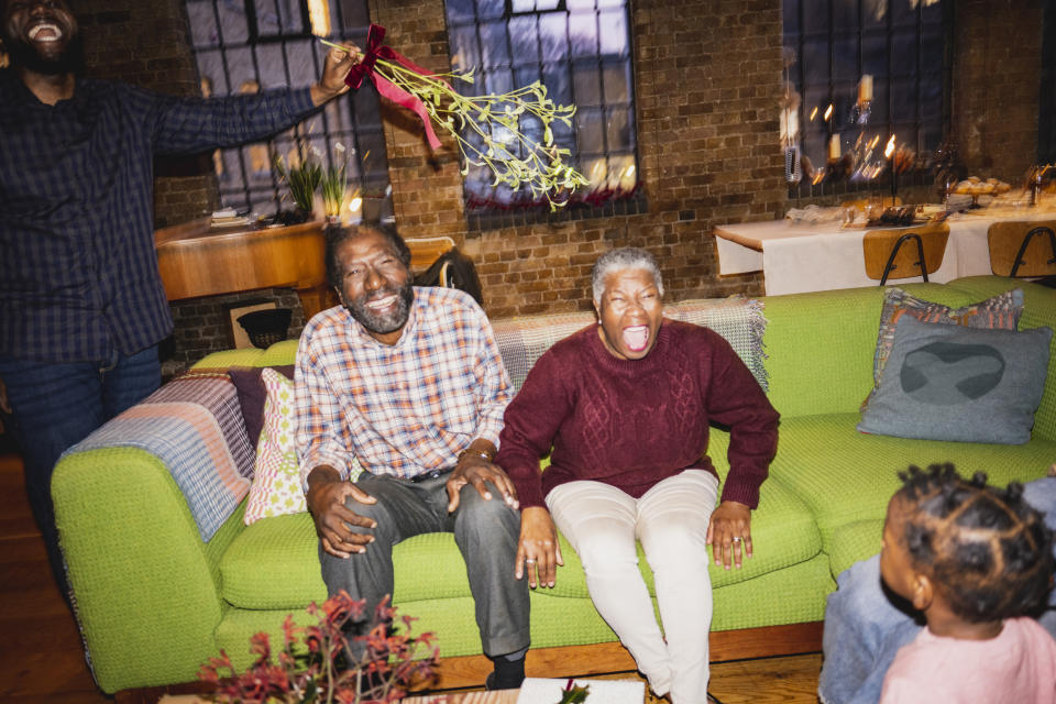 An older couple is surprised and delighted by a person holding mistletoe above them as they sit on a couch. A child looks on