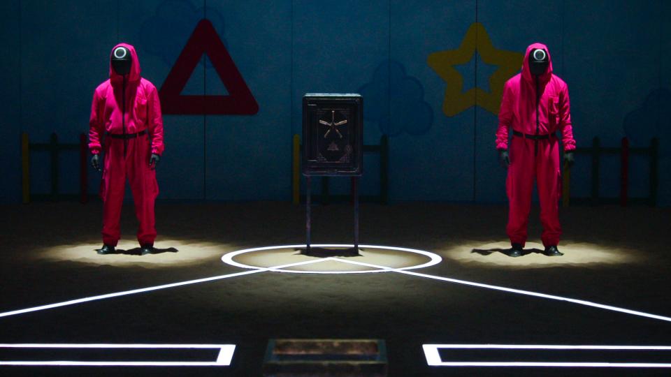 two men in pink jumpsuits and masks stand under spotlights next to a squid game pattern on the floor