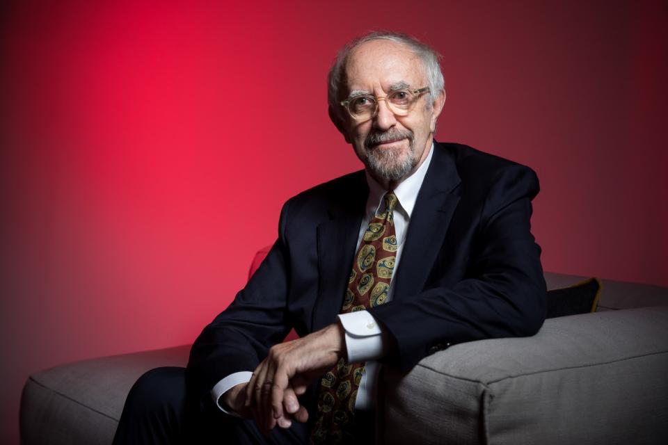 Jonathan Price sits in a gray chair in front of a red backdrop