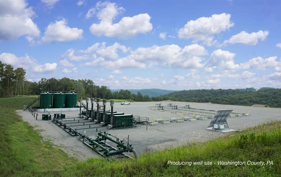 Natural gas well site with pumping and storage equipment, amid a green lawn and forest site under a partly cloudy blue sky.