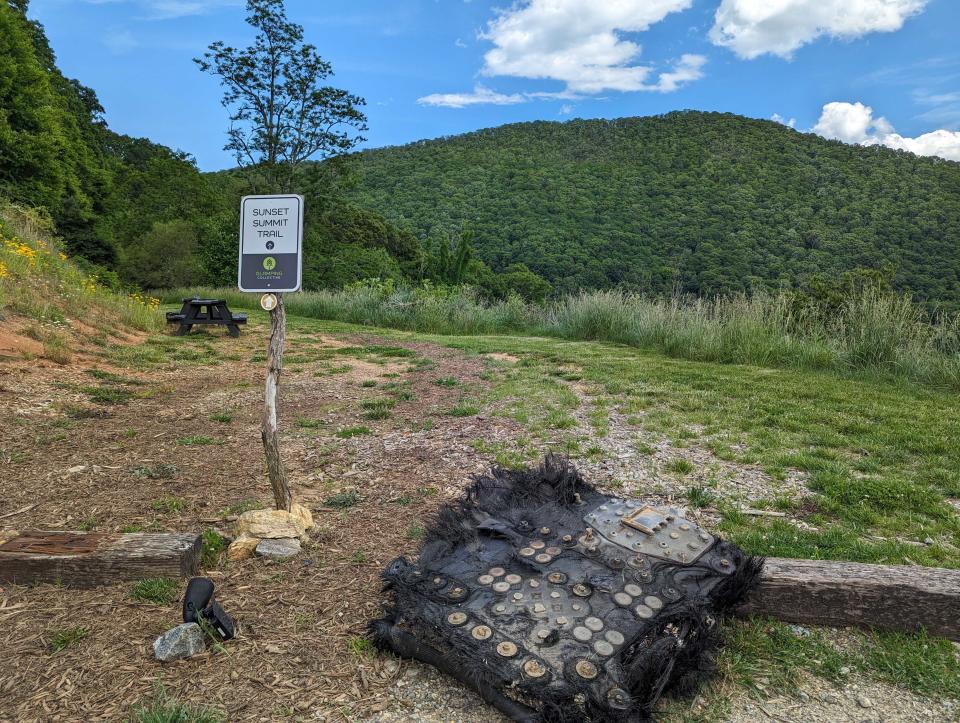 a large black piece of fiberglass covered in metal bolts and plates lies on the ground next to a path leading to a forest.  Mountains can be seen rolling in the distance