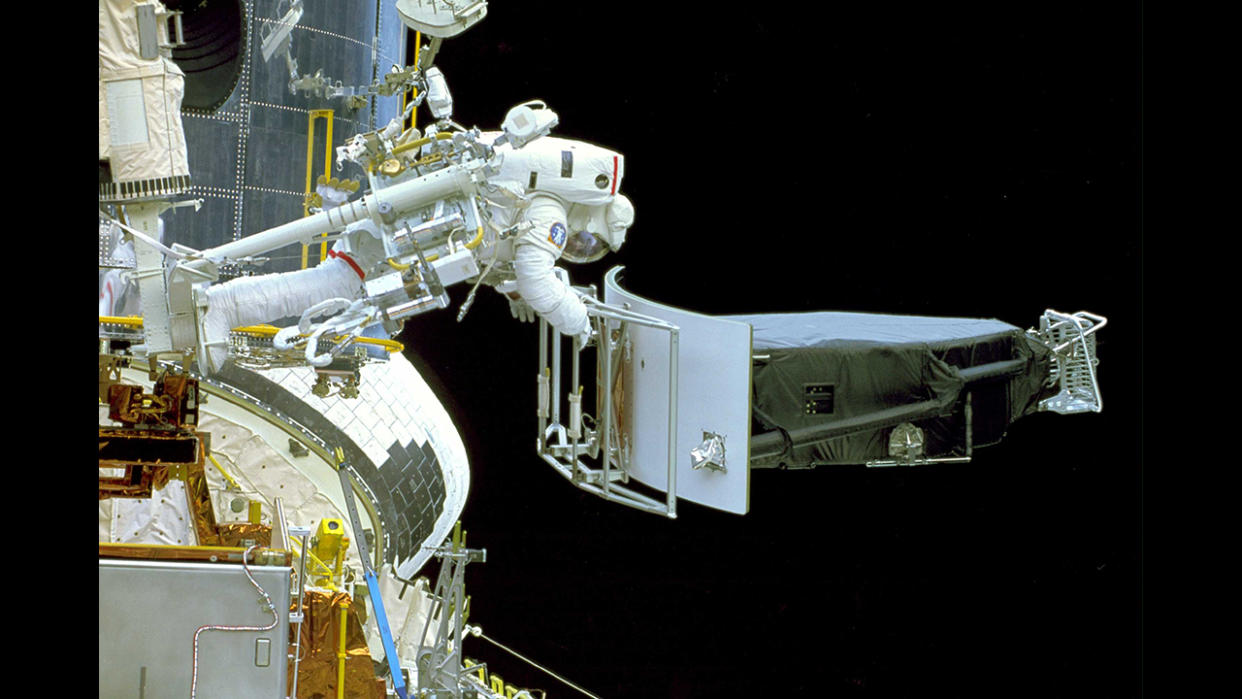  A human in a spacesuit is strapped at the feet to a mechanical arm. the astronaut appears horizontal, facing downward, holding a large space component. 