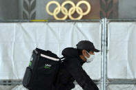 An Uber Eats delivery person carries items near the Japan National Stadium, where opening ceremony and other events are planned for postponed Tokyo 2020 Olympics, with engravings in honor of 1964 Tokyo Olympics seen on the side of the stadium wall behind the fence Tuesday, April 6, 2021, in Tokyo. Two top officials of Japan's ruling LDP party on Thursday, April 15, 2021, said radical changes could be coming to the Tokyo Olympics. One went as far to suggest they still could be canceled, and the other that even if they proceed, it might be without any fans.(AP Photo/Eugene Hoshiko)