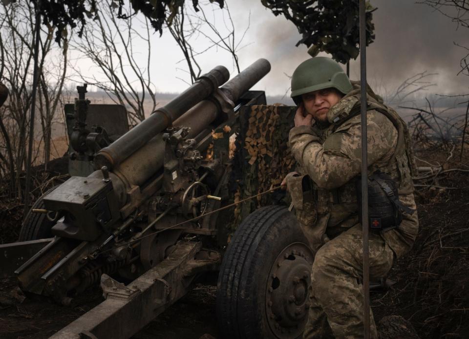 A Ukrainian soldier in combat gear and a helmet squats and covers his ear beside a M101 howitzer