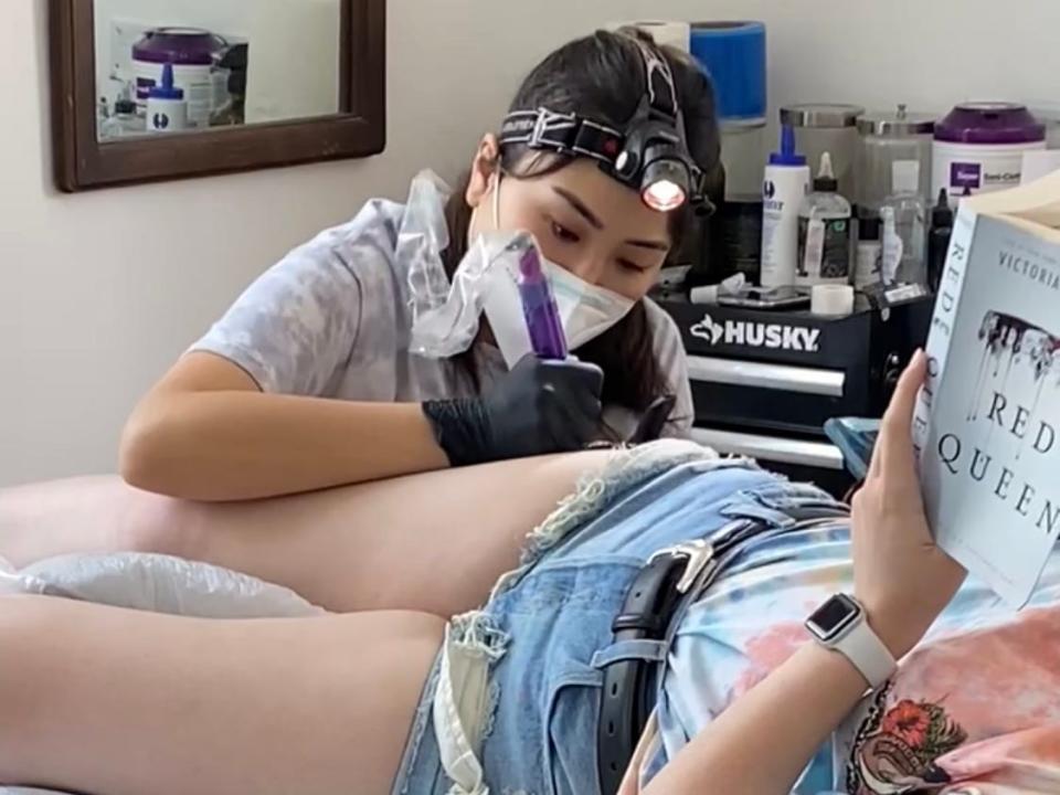 The writer tattooing a client on their leg while the client reads a book