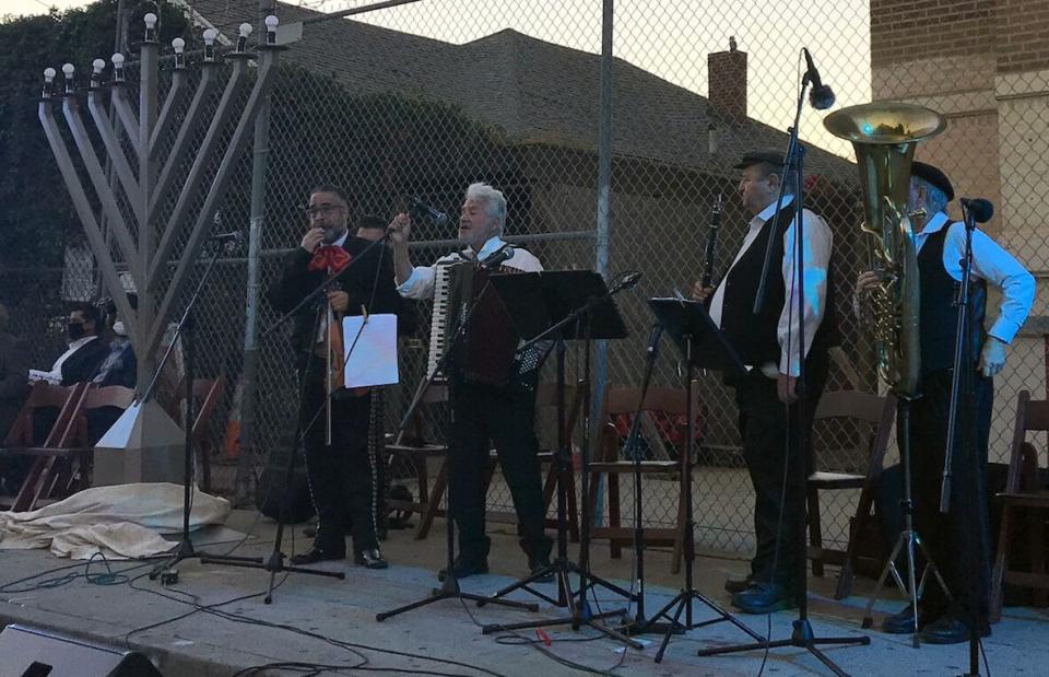 Four men with musical instruments stand beside a giant menorah.