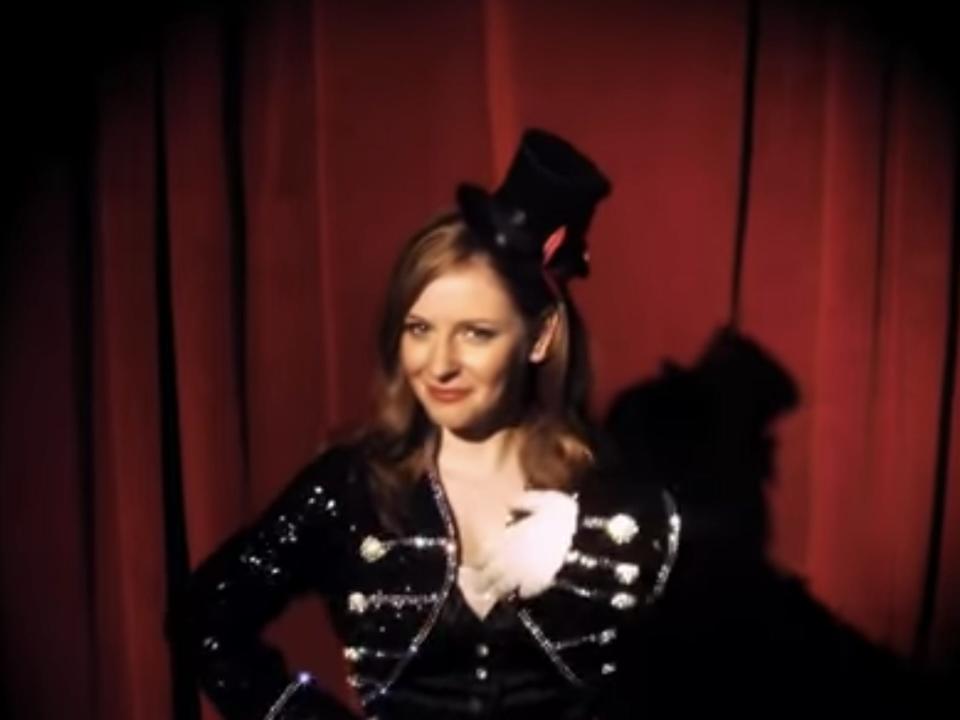 Holly J dressed as a carnival ring leader with a tiny top hat presenting a red-velvet curtain
