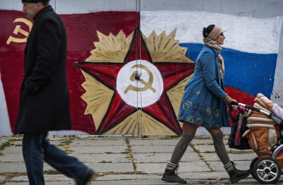 FILE - People pass by the Soviet Union's flag and symbol center, and a Russian flag, right, painted on the gate of a TV company in Luhansk, the territory controlled by pro-Russian militants, eastern, eastern Ukraine, Oct. 27, 2015. Amid fears of a Russian invasion of Ukraine, tensions have also soared in the country’s east, where Ukrainian forces are locked in a nearly eight-year conflict with Russia-backed separatists. A sharp increase in skirmishes on Thursday raised fears that Moscow could use the situation as a pretext for an incursion. (AP Photo/Max Black, File)