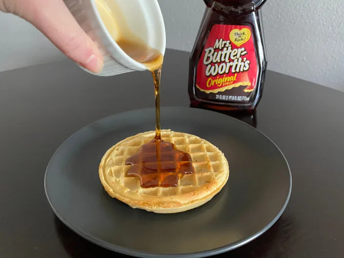 hand pouring white ramekin of mrs butterworth's syrup over a frozen waffle