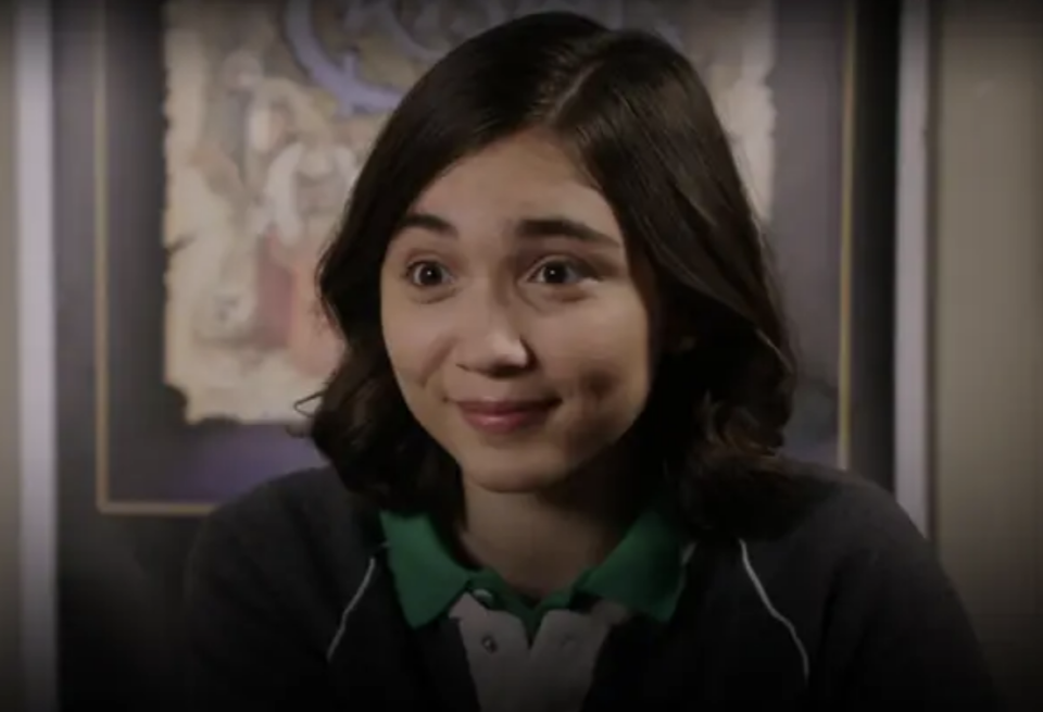 Rowan with shoulder-length dark hair, wearing collared shirt and dark jacket, smiles warmly in front of artwork