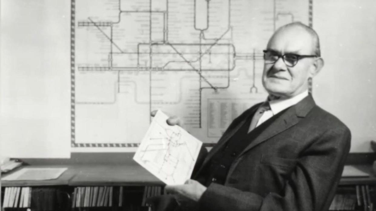 A black and white image of Harry Beck wearing a suit and sitting in front of a London Underground map which is on the wall behind him