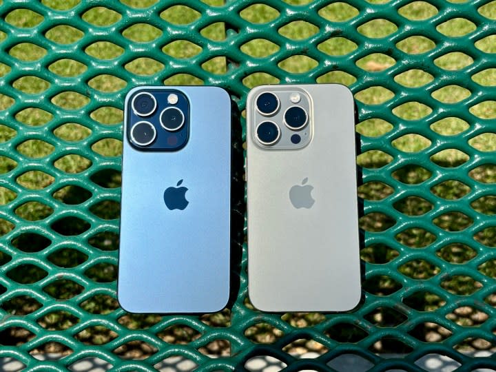 Blue Titanium (left) and Natural Titanium iPhone 15 Pros on a green park bench.