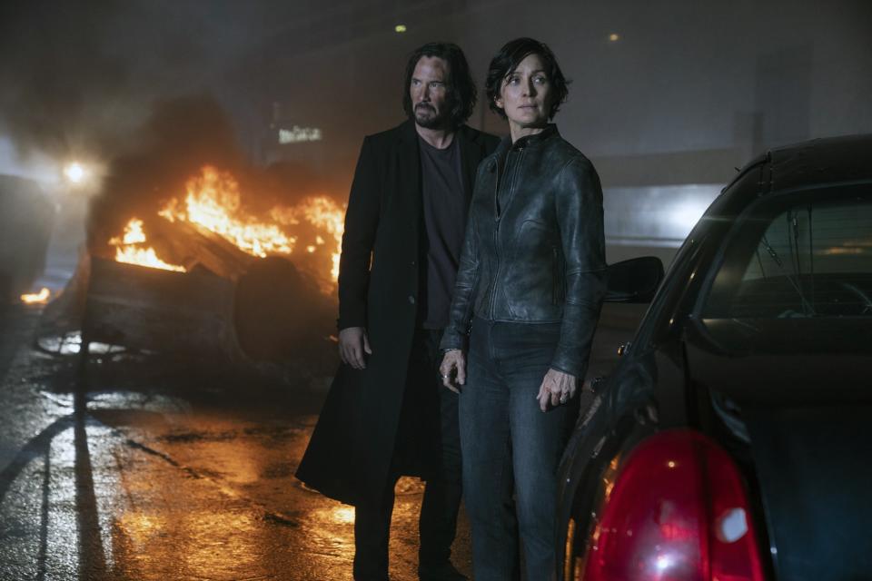 Moss stands next to Keanu Reeves while a car burns in the background