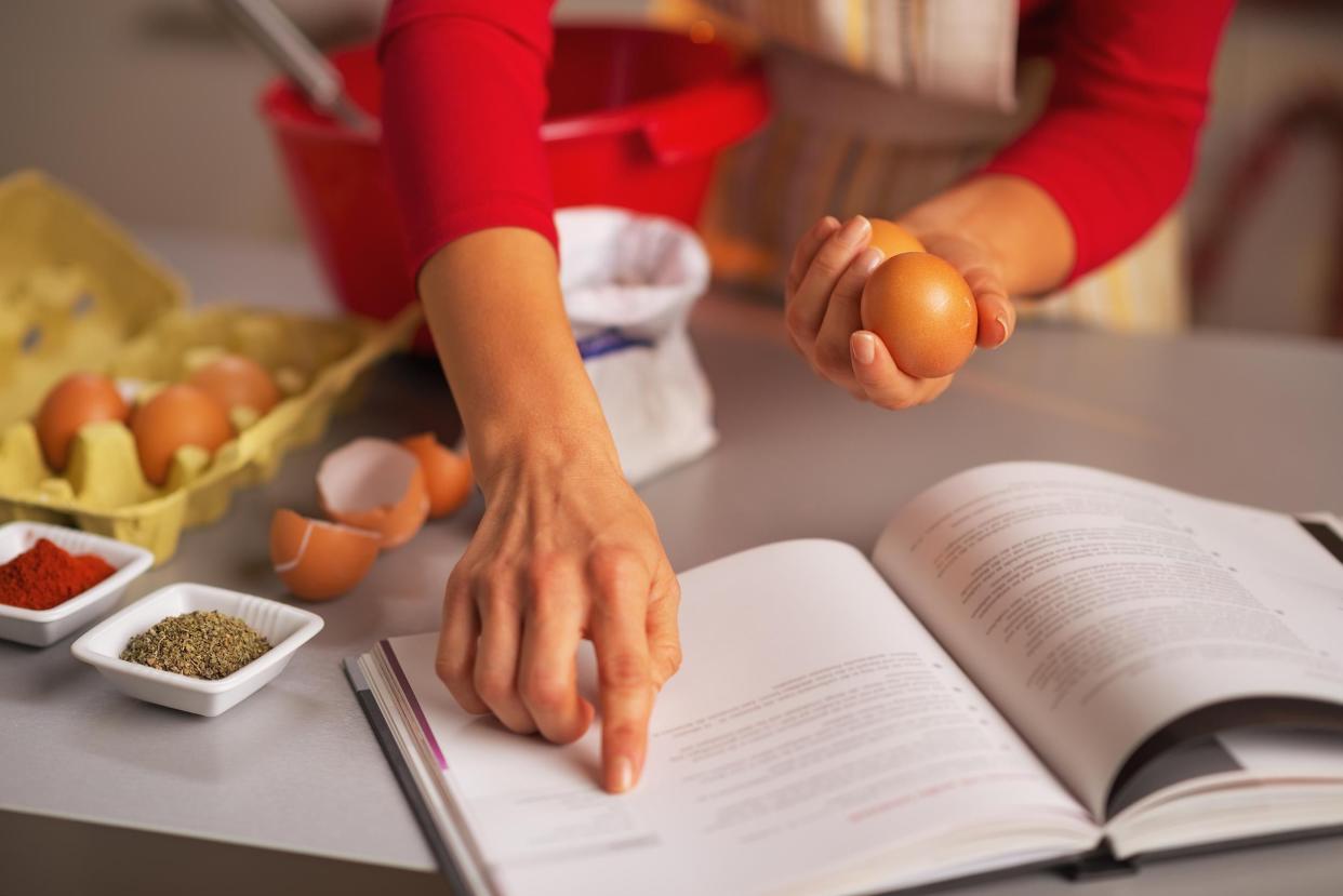 Focus of woman's hand on cookbook as she checks it while holding two eggs, she is in the midst of cooking, kitchen countertop with several ingredients and a big red bowl in the background