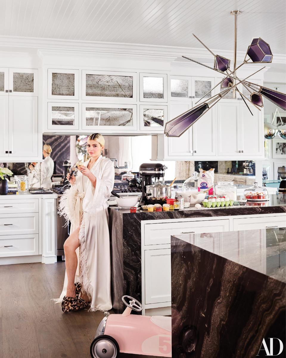 Jenner—wearing a vintage robe, an LPA bodysuit, and Shay Fine Jewelry—in her kitchen. Blown-glass chandelier by Gabriel Scott; custom cabinetry by Martyn Lawrence Bullard.