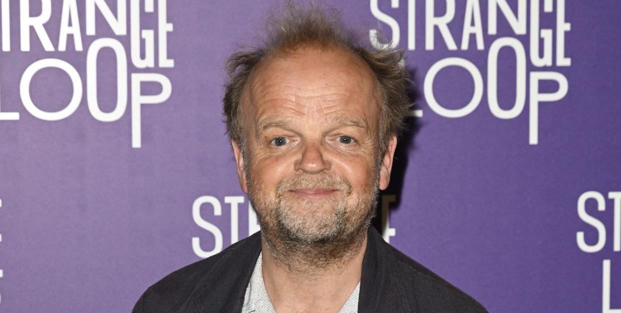 toby jones, an older man stands smiling at the camera, he has brown hair, wears a white shirt with jeans and black jacket