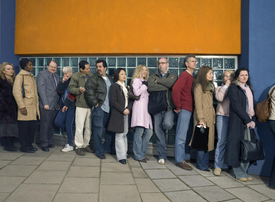 A line of diverse people stand outside, waiting, dressed in various casual outfits, in front of a building