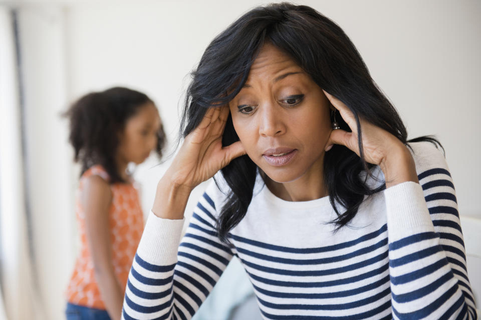 A concerned mother, wearing a striped shirt, rests her head in her hands while a young girl stands in the background