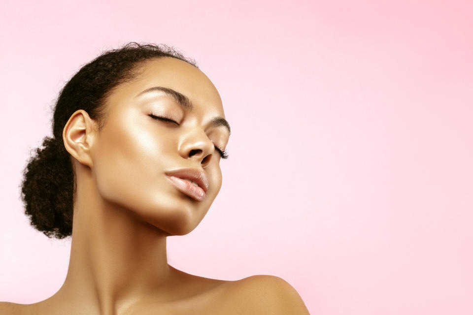 A model with glowing skin and her dark curly hair tied back is seen in three-quarter profile with her eyes closed.