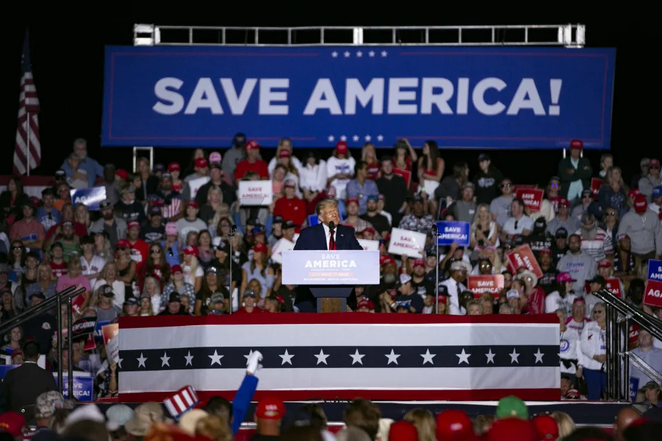 Former President Donald Trump, surrounded by supporters, speaks under a blue banner that says: Save America!