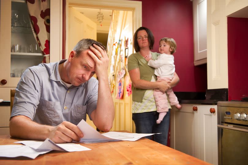 A worried dad reading a letter at the table while mum holds a baby in the kitchen