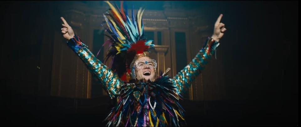 Taron Egerton as Elton John, wearing teal and gold sequinned sleeves with a brightly coloured feathered sequinned body and matching headdress. He has his arms raised and is smiling widely