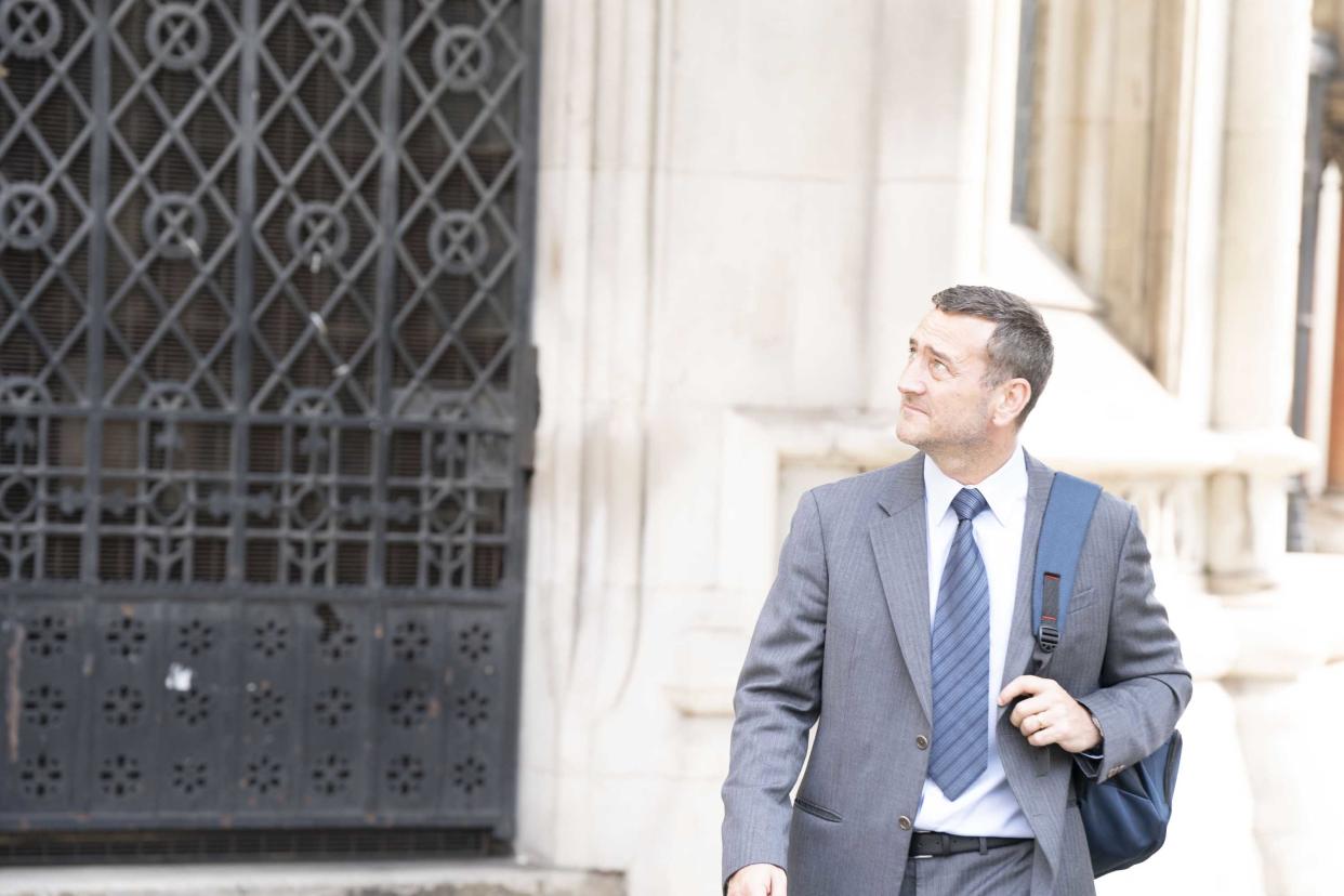 Actor Lee Mellor wearing a grey suit with a blue tie. He has a backpack on one shoulder.