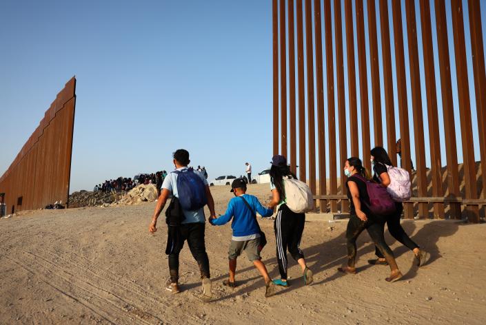 Immigrants cross through a gap in the U.S.-Mexico border barrier as others await processing by the U.S. Border Patrol on May 20, 2022 in Yuma, Ariz.