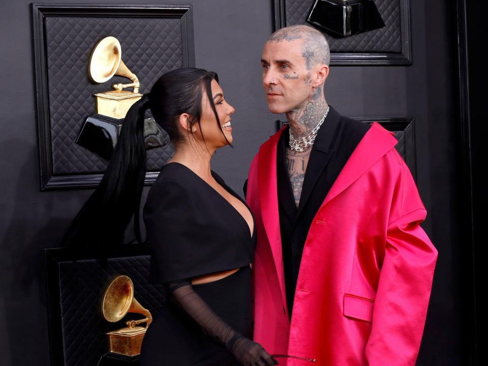 Kourtney in a black long-sleeve jumpsuit. Travis in a neon pink oversized coat over a black suit with no shirt and a crystal necklace.