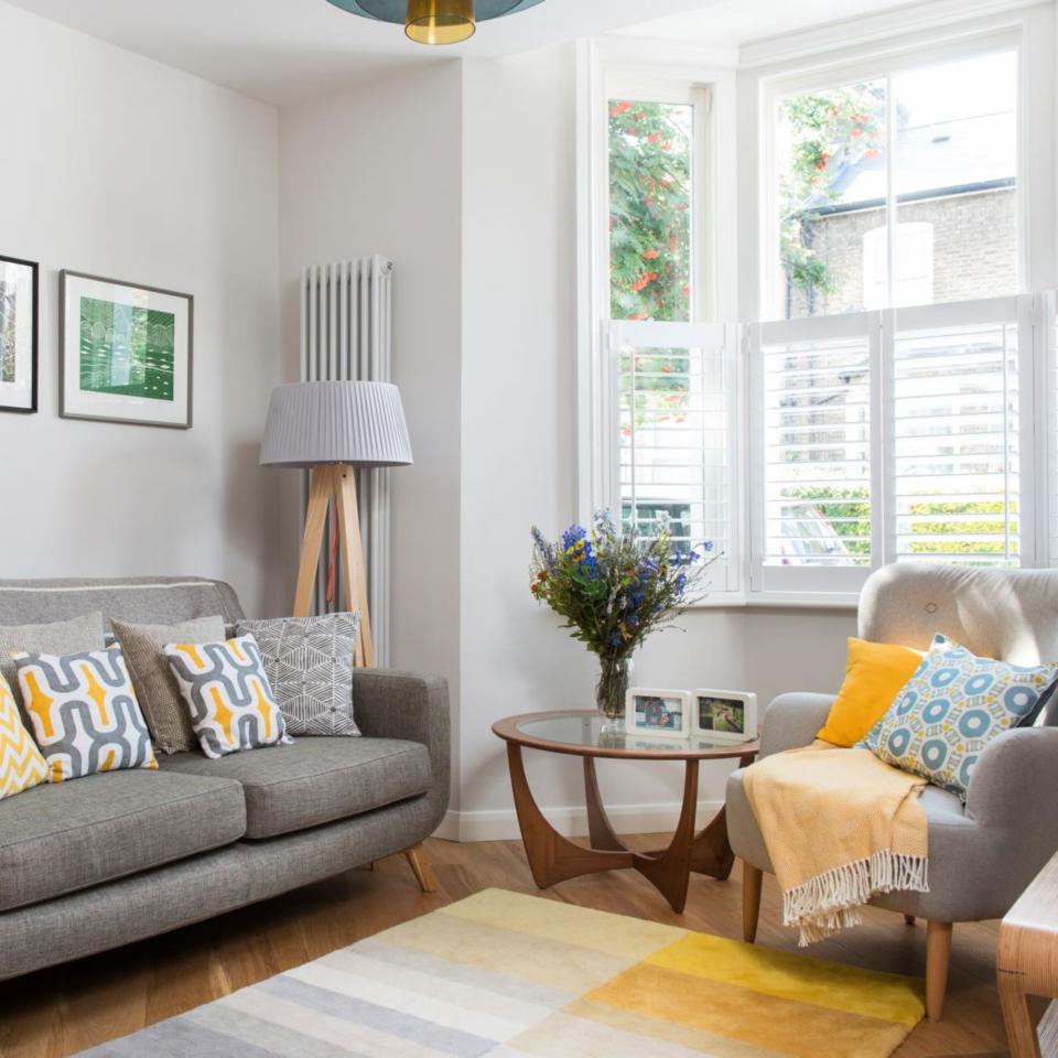 Living room with light grey sofa and armchair with yellow accent cushions and rug, and shutters on window Extended and renovated three bedroom Victorian semi detached house in southeast London, home of Deenie Lee and Martin Thomas