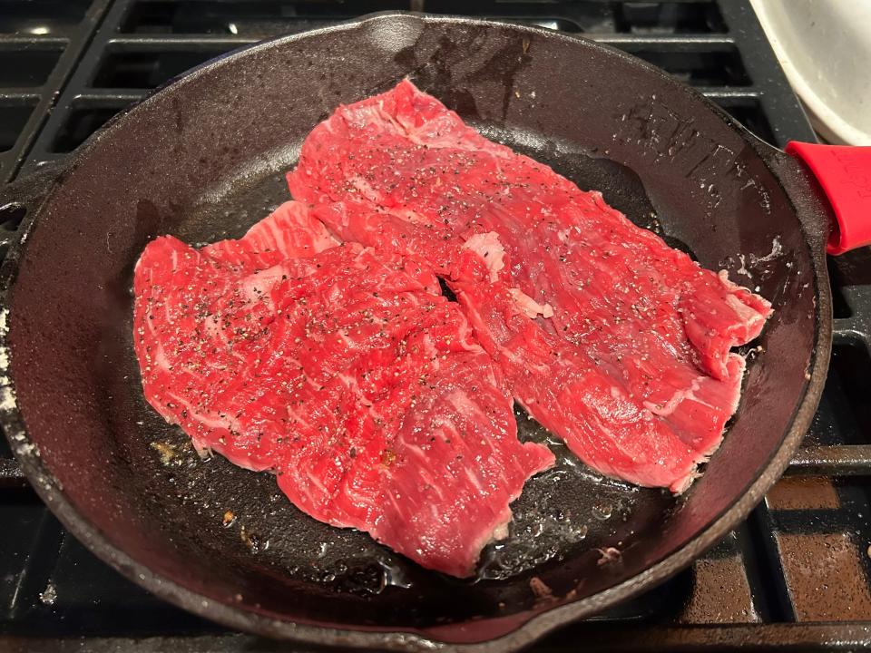 Two pieces of thin flank steak seasoned in a cast-iron skillet on the stove.