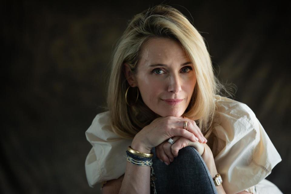 A woman with shoulder length blond hair and a golden bracelet looks directly into the camera.