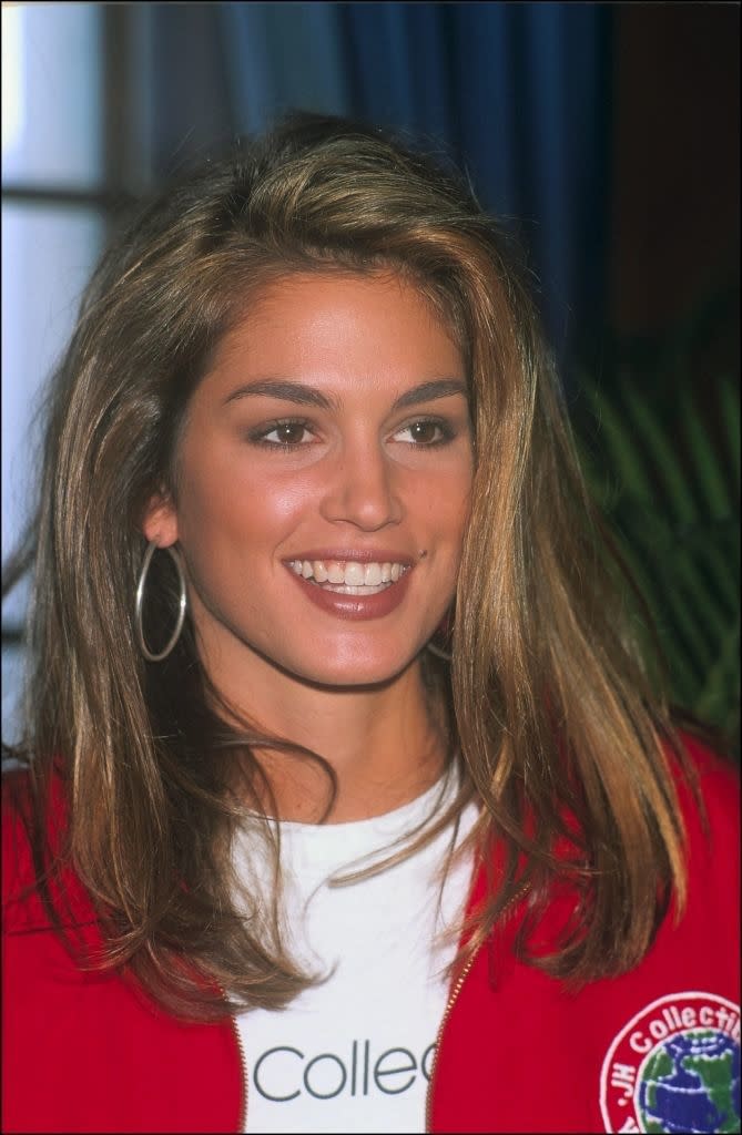 Cindy Crawford with her signature side part, wearing hoop earrings, a white shirt, and a red jacket with a patch