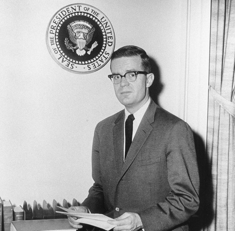 Ted Sorensen poses holding a document in front of a seal hanging on the wall, marked: Seal of the President of the United States.