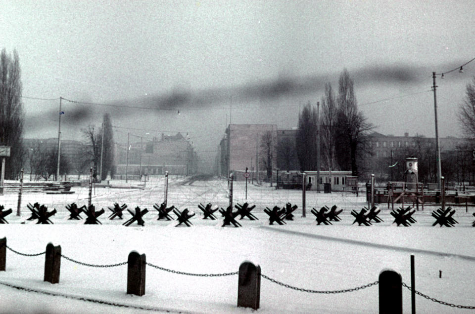 Antitank obstacles line a snowy field in front of large buildings in the distance.