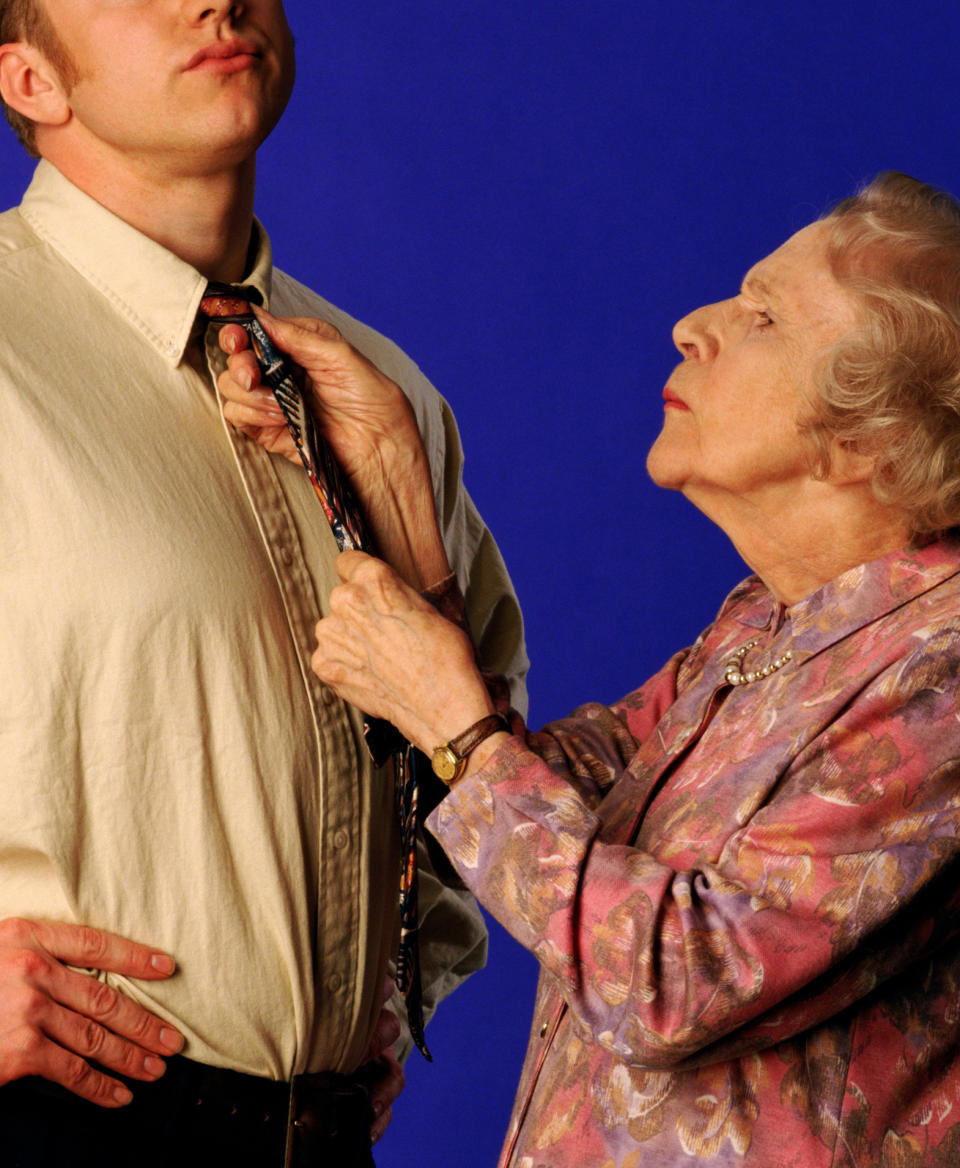An elderly woman adjusts a young man's tie. The scene appears formal, suggesting a wedding or significant event preparation